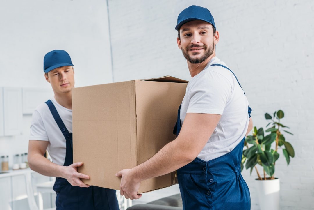 two-handsome-movers-transporting-cardboard-box-in-apartment-e1620131893815.jpg